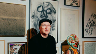 A man wearing a black cap, black shirt, and glasses sits in an art studio surrounded by framed drawings and sculptures. A colorful painted bust sits on a green table to his right.
