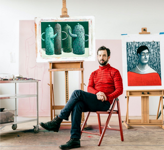 A man in a red knit sweater and dark pants sits on a red chair in an artist studio, surrounded by two easels displaying paintings. One painting shows three greenish-blue abstract forms, and the other shows a portrait with a red top and dark background.