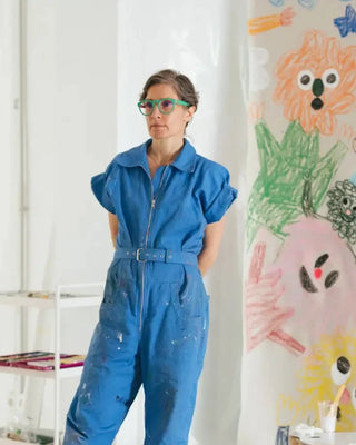 Artist wearing a blue jumpsuit standing in a room with colorful artworks on the walls.