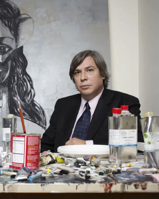 Artist sitting at a table with art supplies and a mural of a face in the background