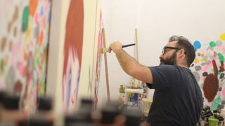 A man wearing glasses and a dark t-shirt paints on a canvas easel in a brightly lit studio filled with colorful artwork.