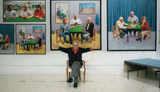 Artist sitting in a chair with paintings of people playing cards on a wall behind him.