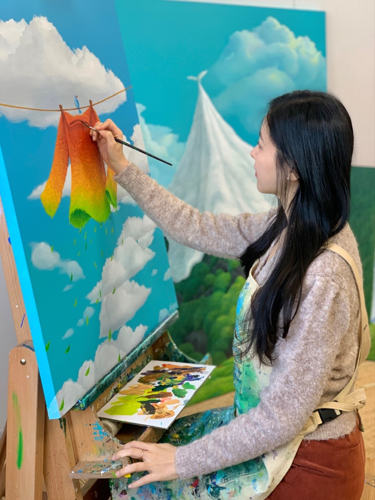 Mulgil Kim, one of the up and coming Contemporary Korean artists to watch, poses in front of her painting of a colourful garment hanging against a cloudy sky.