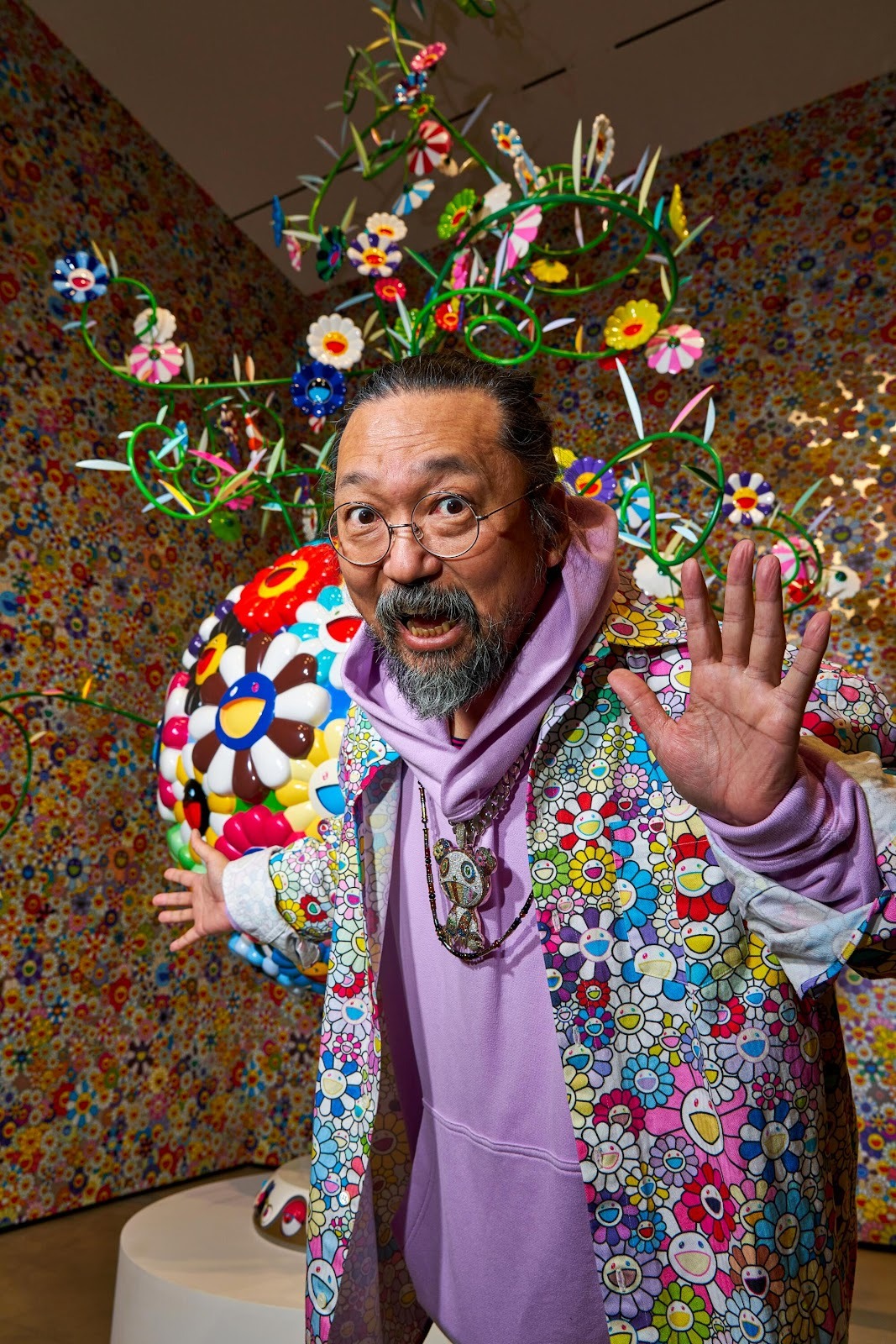 Japanese Contemporary artist Takashi Murakami poses in front of a wall of his smiling flowers, wearing clothing with the same motifs, during his exhibition at the Broad museum.